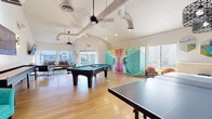 This single-floor amenity space is thoughtfully designed to foster community and recreation, combining energetic game areas with comfortable lounging zones. The game room serves as the centerpiece, featuring soaring vaulted ceilings with exposed ductwork that lends an industrial-modern aesthetic, warm hardwood-style flooring, and an abundance of natural light streaming through large windows. A full-size pool table and ping-pong table anchor the space, while a shuffleboard table paired with a large wall-mounted television creates a dedicated entertainment zone. Colorful, artfully designed package lockers add a vibrant visual focal point to the room, complemented by eclectic wall art and decorative mirrors.

A relaxed lounge area transitions seamlessly from the game room, furnished with a contemporary sofa, accent chairs, and a plush area rug, all positioned to take advantage of views toward the outdoor pool area. Ceiling fans throughout maintain comfort across the open layout. A separate corridor seating nook features bold yellow and pink modular chairs with round side tables, offering a more intimate setting alongside glass-partitioned views into an adjacent meeting or dining area. Direct access to the outdoor pool and surrounding amenities is provided through large glass doors, blurring the line between interior comfort and outdoor recreation.