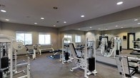 This fitness center is designed to support a well-rounded workout routine within a single-level layout. The space is outfitted with a selection of strength training machines, including a cable/pulley system and a multi-function weight bench, providing users with options for both upper and lower body exercise. A dumbbell rack lines one wall, offering a range of free weights for varied training needs. A rowing machine adds a cardiovascular element to the facility's equipment offerings. Large mirrors span one full wall, enhancing the sense of space and allowing users to monitor their form during exercise. The flooring consists of a durable, low-pile carpet with exercise mats placed beneath key equipment for added cushioning and safety. Recessed lighting is evenly distributed across the ceiling, providing consistent, well-distributed illumination throughout the room. Several windows allow natural light to supplement the artificial lighting, contributing to a bright and energizing atmosphere. The walls are finished in a neutral gray tone, lending the space a clean, modern aesthetic that complements the contemporary fitness equipment. Overall, the facility presents a functional and organized environment suited to a variety of fitness goals.