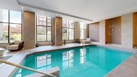 Located at 815 Avenue Plymouth in the prestigious neighbourhood of Mont-Royal, Québec, this upscale indoor pool and amenity facility offers a refined retreat within a contemporary residential setting. The single-level space is generously proportioned, encompassing a substantial floor area designed to promote comfort and relaxation.

At the heart of the facility is a full-length indoor swimming pool with crystal-clear turquoise water, framed by large-format light stone tiles that extend throughout the entire space. Towering floor-to-ceiling windows flank the pool area, flooding the interior with abundant natural light while offering views of the surrounding landscaped grounds and the residential buildings beyond.

Warm-toned stone-clad columns punctuate the poolside, adding an architectural elegance that complements the neutral, spa-like palette of the space. Tasteful lounge seating — including cushioned chaise lounges with marble accent tables — is thoughtfully arranged both poolside and in a dedicated relaxation alcove, creating intimate areas to unwind.

A glass-enclosed entrance vestibule connects the facility to the broader building, featuring additional seating and direct garden-facing double doors that reinforce the seamless indoor-outdoor flow. Recessed lighting throughout ensures a warm, inviting ambiance at any time of day. This facility embodies a calm, resort-inspired atmosphere ideal for leisurely recreation.