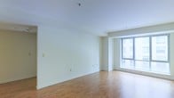 This single-floor space presents a clean, minimalist aesthetic defined by its open-plan living area. The room is finished with smooth light wood flooring that extends throughout, lending warmth and continuity to the interior. Crisp white walls and a white ceiling create a bright, airy atmosphere that feels open and versatile.

A standout feature is the large floor-to-ceiling window positioned along one wall, framing views of the surrounding urban landscape and drawing in generous amounts of natural light. The window's substantial size ensures the space feels expansive and well-illuminated throughout the day.

The room's layout is straightforward and flexible, offering a blank canvas suitable for a variety of furniture arrangements and uses. Subtle architectural detailing, including a recessed ceiling beam near the window wall, adds a touch of structure to the otherwise clean design. Electrical outlets are thoughtfully distributed along the baseboards, and a fire sprinkler is visible in the ceiling, reflecting modern safety standards. Overall, the space conveys a contemporary, functional character with an emphasis on light, simplicity, and adaptability.