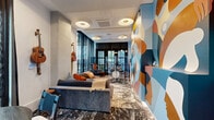 Located in the heart of Nashville at 900 Second Ave N, this thoughtfully designed music-inspired space captures the creative spirit of Music City. Floor-to-ceiling sheer black curtains frame expansive windows that flood the interior with natural light and offer views of the surrounding greenery, creating a dynamic interplay between the indoors and outside environment. A full Ludwig drum kit sits behind a clear acrylic sound shield, accompanied by a professional mixing console and keyboard workstation, making the space fully equipped for musical performance and production. A Taylor acoustic guitar is mounted on the wall as both a functional instrument and a design statement. The lounge area features a plush gray sofa accented with warm-toned throw pillows and a richly textured area rug, providing a comfortable setting for collaboration or relaxation. A round dining table with chairs offers an additional gathering spot within the space. The most striking visual element is a large-scale, bold mural covering an entire wall, depicting abstract human figures in a vibrant palette of blue, orange, terracotta, and cream — lending an unmistakable artistic identity to the room. The overall ambiance blends the warmth of a creative studio with the curated aesthetic of a boutique hospitality setting.