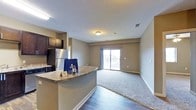 This single-floor residential unit offers a thoughtfully designed open-concept layout that seamlessly connects the kitchen and living areas. The kitchen is equipped with dark cabinetry, stainless steel appliances, and a granite-topped breakfast bar that provides both additional counter space and a casual dining option. The transition from kitchen to living area is marked by a shift from rich hardwood-style flooring to plush carpeting, creating a warm and inviting atmosphere throughout.

The living area is generously proportioned, offering ample room for a variety of furniture arrangements. Neutral, warm-toned walls complement the overall palette of the space, contributing to a clean and contemporary aesthetic. A textured ceiling adds subtle visual interest overhead, while flush-mount lighting provides consistent illumination.

Natural light is a standout feature of the living area, which opens directly onto a private balcony through a large sliding glass door. The balcony offers outdoor views of the surrounding area, making it an ideal spot for relaxation. Visible HVAC vents are integrated into the ceiling, ensuring year-round comfort. Overall, the space combines practical functionality with comfortable, modern living in a cohesive and well-appointed design.