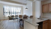This single-floor residence is designed with an open-concept layout that seamlessly connects the kitchen, dining, and living areas into one cohesive, flowing space. The kitchen features a striking granite countertop island with seating, offering both a functional workspace and a casual gathering spot. A pendant light fixture hangs above, adding a contemporary touch to the space.

The dining area sits at the heart of the open floor plan, anchored by a dark-framed table with upholstered seating, bridging the kitchen and living room with ease. Hardwood flooring in a warm, natural tone runs throughout, lending warmth and continuity to the entire space.

The living room is bathed in natural light, courtesy of a generous bank of windows framed by sheer white curtains. The soft, diffused light enhances the airy ambiance of the room, which is furnished with a tufted sofa and coordinating armchairs arranged around a glass-top coffee table. The neutral color palette of whites, beiges, and grays creates a calm and inviting atmosphere throughout. Structural beams add a subtle architectural detail overhead, contributing character and depth to the overall design. The space is thoughtfully arranged to maximize both comfort and functionality.