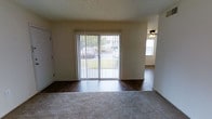 This single-floor residential space offers a practical and straightforward layout designed for comfortable everyday living. The main living area is a welcoming room featuring a combination of plush carpeting and dark hardwood-style flooring, creating a clear distinction between relaxation and transitional zones. A set of sliding glass doors dressed with vertical blinds anchors the room, allowing natural light to filter in while offering a view of the surrounding greenery outdoors. The textured ceiling adds a classic residential character to the space. Adjacent to the living area, a connected room is visible, outfitted with a ceiling fan for added comfort and continuing the hardwood-style flooring throughout. The transition between rooms is open and fluid, contributing to a sense of continuity across the floor plan. The neutral wall tones throughout provide a clean, versatile backdrop that can accommodate a variety of interior styles. Overall, this single-level space combines functional design with natural light and outdoor connectivity, making it a practical and adaptable living environment.