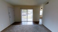 This single-floor residential space offers a practical and straightforward layout designed for comfortable everyday living. The main living area is a welcoming room featuring a combination of plush carpeting and dark hardwood-style flooring, creating a clear distinction between relaxation and transitional zones. A set of sliding glass doors dressed with vertical blinds anchors the room, allowing natural light to filter in while offering a view of the surrounding greenery outdoors. The textured ceiling adds a classic residential character to the space. Adjacent to the living area, a connected room is visible, outfitted with a ceiling fan for added comfort and continuing the hardwood-style flooring throughout. The transition between rooms is open and fluid, contributing to a sense of continuity across the floor plan. The neutral wall tones throughout provide a clean, versatile backdrop that can accommodate a variety of interior styles. Overall, this single-level space combines functional design with natural light and outdoor connectivity, making it a practical and adaptable living environment.