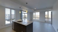 This contemporary single-floor space offers a thoughtfully designed open-concept layout that seamlessly connects the kitchen and living areas. The kitchen is anchored by a striking island with a crisp white countertop and dark cabinetry, complemented by an undermount sink and a modern faucet. Two pendant lights with warm-toned cylindrical shades hang above the island, providing focused task lighting and a stylish focal point. Stainless steel appliances add a polished, functional touch to the culinary space. The open living area benefits from abundant natural light streaming through multiple large windows fitted with horizontal blinds, offering views of the surrounding neighborhood. Sliding glass doors lead directly to an outdoor balcony, further enhancing the sense of openness and bringing the outdoors in. The neutral, light-toned flooring flows continuously throughout the space, creating a cohesive and airy atmosphere. Recessed lighting in the ceiling supplements the natural light, ensuring the interior remains bright and welcoming at any time of day. The clean lines, neutral color palette, and thoughtful layout make this a versatile and modern living environment.