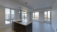 This contemporary single-floor space offers a thoughtfully designed open-concept layout that seamlessly connects the kitchen and living areas. The kitchen is anchored by a striking island with a crisp white countertop and dark cabinetry, complemented by an undermount sink and a modern faucet. Two pendant lights with warm-toned cylindrical shades hang above the island, providing focused task lighting and a stylish focal point. Stainless steel appliances add a polished, functional touch to the culinary space. The open living area benefits from abundant natural light streaming through multiple large windows fitted with horizontal blinds, offering views of the surrounding neighborhood. Sliding glass doors lead directly to an outdoor balcony, further enhancing the sense of openness and bringing the outdoors in. The neutral, light-toned flooring flows continuously throughout the space, creating a cohesive and airy atmosphere. Recessed lighting in the ceiling supplements the natural light, ensuring the interior remains bright and welcoming at any time of day. The clean lines, neutral color palette, and thoughtful layout make this a versatile and modern living environment.