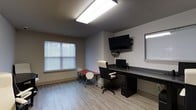 This single-floor office space is thoughtfully designed to support a productive working environment. The main area features a built-in desk that runs along one wall, providing generous workspace and accommodating a desktop computer setup. A wall-mounted flat-screen television is positioned above the desk, complemented by an additional shelf for media equipment, making the space well-suited for presentations or collaborative viewing. A comfortable ergonomic office chair completes the primary workstation. A smaller folding table with two chairs offers an additional space for meetings or collaborative work, adding flexibility to the layout. The room is finished with light wood-look flooring that lends a clean, modern aesthetic throughout. Neutral-toned walls keep the space feeling open and uncluttered. Natural light filters in through multiple windows fitted with horizontal blinds, allowing for adjustable brightness throughout the day. Overhead, a large recessed fluorescent light fixture ensures consistent and bright illumination regardless of outside conditions. Overall, the space strikes a balance between simplicity and functionality, providing a well-lit, organized environment suited for focused work, small meetings, or administrative tasks.
