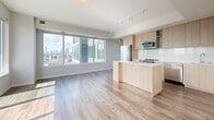 This contemporary high-rise unit offers a well-designed single-floor layout with 3 bedrooms and 3 bathrooms. The open-concept kitchen and living area serves as the heart of the space, featuring light wood cabinetry, a central island with a white countertop, stainless steel appliances including a refrigerator and built-in microwave, and track lighting overhead. Large floor-to-ceiling windows flood the combined living and kitchen area with abundant natural light while framing impressive panoramic city skyline views. Hardwood flooring in warm natural tones runs consistently throughout the unit, lending cohesion and warmth to every room. A wide hallway connects the living areas to the three bedrooms, each of which benefits from generous windows offering elevated cityscape views. The primary bedroom is a comfortable, well-proportioned retreat with access to its own bathroom. The bathrooms are finished in a clean, modern style with white subway-tiled shower-tub combinations, light wood vanities, and large-format tile flooring. One bathroom also features a built-in shelving closet for additional storage. A dedicated closet provides further organizational space. The overall aesthetic is clean and minimalist, with white walls, crisp trim, and contemporary fixtures creating a bright, cohesive living environment throughout.