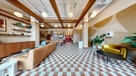 This thoughtfully designed single-floor commercial space offers a generous footprint that balances open communal areas with private workspaces. The hallway serves as the heart of the space, immediately drawing attention with its striking tri-color checkered tile floor in terracotta, gray, and white — a bold mid-century modern design statement. Exposed wood beam ceilings add architectural warmth, while globe pendant lights provide soft, ambient illumination throughout. Large floor-to-ceiling windows flood the entrance area with natural light and offer views of the surrounding streetscape. A built-in reception desk with open shelving, decorative plants, and curated accessories creates a polished, welcoming atmosphere. An exposed brick accent wall adorned with abstract murals adds artistic character, complemented by warm wood paneling throughout. A kitchenette bar area with counter seating is ideal for casual gatherings or refreshments. The hallway corridor, accented with vibrant neon signage, leads to the private office — a focused meeting room featuring deep red carpet, a round conference table, wood chairs, and a wall-mounted display screen, perfectly suited for small group meetings or collaborative work sessions.