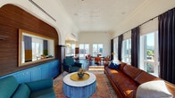 Located within The Hoxton Hotel on NW Quimby Street in Portland, Oregon, this single-floor suite offers a thoughtfully curated event and gathering space with a distinctive blend of classic architectural details and contemporary design. The space is defined by its striking embossed tin ceilings, generous ceiling height, and rich herringbone-patterned hardwood floors that flow throughout.

The main lounge area is anchored by a substantial leather sofa, teal upholstered seating, and a marble-topped round coffee table set atop a traditional area rug. An arched wood-slatted feature wall with a gold-framed mirror adds warmth and visual depth near the entrance. Large windows and a sliding glass door flood the space with natural light and open to an outdoor terrace, revealing sweeping views of the Portland skyline and surrounding greenery.

A separate dining and meeting area accommodates a round table with leather chairs, positioned to take full advantage of panoramic city views through floor-to-ceiling windows dressed with dramatic navy drapes.

The kitchenette is outfitted with bold cobalt-blue cabinetry, brass hardware, white quartz countertops, open shelving with gold brackets, and arched architectural details — combining functionality with elegant design throughout.