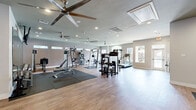 This generously sized fitness center offers a comprehensive workout environment thoughtfully designed to accommodate a wide range of exercise needs. The space features durable hardwood-style flooring throughout, complemented by protective rubber mat sections strategically placed beneath weight and strength equipment. Multiple large ceiling fans are installed across the ceiling, ensuring consistent airflow and comfort during workouts, alongside recessed lighting that keeps the space bright and well-illuminated.

The main workout area is extensively equipped with a smith machine, cable cross machines, a multi-station strength system, adjustable benches, a full dumbbell rack with weights ranging across multiple sizes, kettlebells, medicine balls, and a wall-mounted television for entertainment. A dedicated cardio zone near the arched windows houses treadmills, an elliptical, and a stationary bike, all benefiting from natural light and outdoor views through the large windows and glass-paneled door.

A secondary alcove area provides additional training space with more cardio equipment and a stability ball, along with a built-in water fountain for convenient hydration. The neutral-toned walls, clean layout, and open floor plan contribute to a professional and welcoming fitness environment suitable for a full-body training regimen.