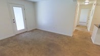 This single-floor residential space offers a clean, functional layout designed for comfortable everyday living. The interior is characterized by neutral white walls and warm beige wall-to-wall carpeting that runs throughout, creating a cohesive and understated aesthetic. The main living area is generously proportioned, providing ample room for furniture arrangement and open movement. White baseboards provide a crisp contrast against the soft carpeting, adding a touch of polished detail to the space. Natural light enters through what appears to be a glass door or large window on one side, brightening the room and contributing to an airy, welcoming atmosphere. A hallway visible toward the rear of the space suggests additional rooms beyond, offering practical connectivity throughout the home. Electrical outlets are thoughtfully placed along the walls for versatile use of the space. The overall ambiance is calm and move-in ready, with a blank-canvas quality that makes it easy to personalize. The consistent flooring and color palette throughout lend a sense of openness and continuity, making this a straightforward and adaptable single-floor living environment.
