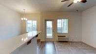 This single-floor space showcases an open-concept layout that seamlessly connects the kitchen and living areas. The kitchen features a sleek white countertop with an overhang suitable for seating, complemented by a pendant chandelier that adds a touch of elegance. The living area transitions from hardwood-style flooring to carpeting, creating distinct zones within the open floor plan. Multiple windows, including a glass-paneled exterior door, flood the space with abundant natural light while providing views of the surrounding greenery. A ceiling fan in the living area ensures year-round comfort, while a wall-mounted PTAC unit offers additional climate control. The neutral, warm-toned walls contribute to a clean and welcoming ambiance throughout. The layout is practical and efficient, making excellent use of the available space while maintaining an airy, open feel. The combination of modern finishes, natural light, and functional design elements makes this a comfortable and versatile living environment.