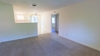 This single-floor space features an open layout that connects the main living area to the kitchen and beyond. The living room is finished with neutral-toned carpet, white walls, and clean white baseboards, giving the space a bright and simple aesthetic. A textured ceiling with a central HVAC vent adds a functional overhead element, while a ceiling fan in the adjacent kitchen area aids in air circulation. The kitchen is partially visible behind a half-wall partition that creates a natural divide while maintaining an open feel. White cabinetry lines the kitchen walls, and tile or hard flooring distinguishes it from the carpeted living area. Natural light filters in through multiple windows, including one visible in the kitchen and another in a hallway beyond the living room. A doorway leads to an additional room or hallway, suggesting further living space beyond the main area. The overall ambiance is clean, minimalist, and move-in ready, with a neutral palette that lends itself well to a variety of interior styles.