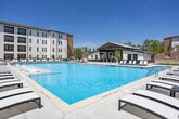 Located at 1801 Riverside Parkway in Douglasville, Georgia, this expansive outdoor amenity space is designed to serve as a central social hub. The centerpiece is a large, resort-style swimming pool with crystal-clear water, surrounded by generous concrete pool decking lined with rows of chaise lounges and shaded umbrella seating, ideal for relaxing under the Georgia sun. The multi-story residential buildings encircle the courtyard on multiple sides, creating a sense of enclosure while maintaining openness to the sky.

Adjacent to the pool is a standalone covered pavilion featuring an exposed wood-beam gabled roof, ceiling fans, and flexible lounge and dining furniture arranged beneath, providing a shaded outdoor gathering area. The pavilion's board-and-batten exterior is complemented by large windows that overlook the pool deck.

Beyond the pool area, the courtyard extends into well-maintained open lawn areas with lush green turf, concrete pathways, and additional seating zones. String lights strung between wooden posts add an inviting ambiance for evening gatherings. A dedicated activity court and landscaped areas with stone detailing round out this thoughtfully designed outdoor community space, offering a variety of amenities for recreation and relaxation.
