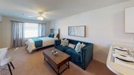 This single-floor studio offers a thoughtfully arranged open-concept layout that maximizes every inch of available space. The kitchen is positioned near the entry and features white cabinetry, a coil-top electric range, a full-size refrigerator, and a counter with warm wood-toned accents beneath. Light tile flooring runs through this area, extending to the front entry door, which adds a clean, practical touch to the arrival experience. The bathroom is efficiently designed with a tub-shower combination, a wide vanity with a white countertop, warm wood cabinetry below, and a large mirror framed by overhead lighting — creating a bright and functional space. The main living area transitions seamlessly from kitchen to living and sleeping zones, all unified by soft, neutral carpeting underfoot. A ceiling fan centered overhead provides both light and airflow throughout. A mirrored sliding-door closet along one wall adds depth and additional storage, visually expanding the room. A window allows natural light to filter in, brightening the space. The open floor plan allows flexible furniture arrangements, accommodating a full sleeping area alongside a living setup with ease, making the studio both practical and comfortable for everyday living.
