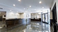 The lobby of Plaza 53 presents a polished and professional atmosphere, immediately establishing a sense of sophistication upon entry. The expansive space is anchored by striking, dark-toned marble floors with inlaid geometric detailing that reflects the overhead recessed lighting, adding depth and visual interest underfoot. A substantial reception desk with dark wood paneling serves as the focal point, positioned prominently to greet visitors and occupants alike. The "Plaza 53" signage is displayed in metallic lettering on the wall, reinforcing the building's identity. A decorative round mirror adorns the white wall behind the desk, complementing the refined aesthetic. Adjacent to the reception area, a secondary service counter with dark wood wainscoting provides additional functionality. The lobby benefits from bright, recessed ceiling lighting distributed evenly throughout, maintaining a well-lit and welcoming environment. Floor-to-ceiling glass entrance doors allow natural light to filter in from the exterior, connecting the interior to the outside. The overall design balances elegance with practicality, making this lobby a functional and visually impressive entry point for the building.