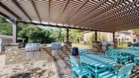 Located at 4950 US-90 W in Mobile, Alabama, this outdoor community amenity space offers a well-designed gathering area tailored for relaxation and social enjoyment. The centerpiece of the space is a substantial wood pergola that provides partial shade over a defined outdoor living area, complete with a stone-clad outdoor kitchen and bar, ideal for entertaining and communal dining. Teal Adirondack chairs add a pop of color and casual seating throughout the paved areas.

The expansive paver patio, constructed with interlocking stone in a varied pattern, creates a durable and visually appealing surface that connects the various functional zones. A stone fire pit area anchors one side of the patio, offering a cozy focal point for cooler evenings. The surrounding landscape features well-maintained lawns, mulched planting beds, young ornamental trees, and mature pines and hardwoods along the perimeter, providing a natural backdrop and sense of privacy.

Brick masonry detailing on adjacent structures complements the stone elements within the amenity area, lending architectural cohesion to the overall design. The space is enclosed by black iron fencing, clearly defining the area while maintaining open sightlines to the surrounding greenery. Overall, this outdoor space delivers a welcoming and functional environment suited for community gatherings.
