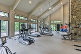 Located at 3000 Cumberland Club Dr in Atlanta, Georgia, this fitness center offers a well-appointed, single-floor workout facility designed with both function and aesthetics in mind. The space is defined by soaring vaulted ceilings with exposed wood beam detailing and white shiplap paneling, creating an open, airy atmosphere that feels expansive and inviting. Recessed lighting complements the abundant natural light that pours in through large floor-to-ceiling windows and glass doors lining multiple walls, offering views of the surrounding lush greenery. The cardio zone is generously equipped with True-brand treadmills, elliptical trainers, a stationary bike, and a rowing machine, all positioned to take advantage of the scenic outdoor views. A dedicated strength and functional training area features a power tower, adjustable bench, and a full rack of medicine balls and stability balls in various sizes. A standout architectural feature is the floor-to-ceiling natural stone fireplace, which anchors one end of the room and adds a warm, rustic contrast to the otherwise modern fitness environment. Direct access to an outdoor patio is available through the glass doors, further enhancing the connection to the natural surroundings. Durable, neutral-toned flooring throughout completes this thoughtfully designed fitness space.