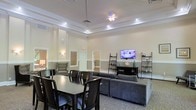 This single-floor space offers an open-plan layout that seamlessly combines a dining area and a lounge, creating a versatile and welcoming environment. The room is anchored by a striking tray ceiling accented with recessed lighting and a central ceiling fan, adding both architectural interest and practical illumination. The walls feature a two-tone finish, lending a refined, classic aesthetic throughout.

The dining area is furnished with a dark wood table and matching chairs, providing ample seating for group gatherings. Adjacent to it, the lounge area features a generously sized gray sectional sofa oriented toward a wall-mounted flat-screen television, supported by lean ladder-style shelving units that offer both display and storage. Wall sconces add warm, ambient lighting along the perimeter.

A carpeted floor in the lounge area transitions the space into a comfortable setting, while an additional seating nook near the windows allows for quieter, more intimate use. Natural light filters in through multiple windows fitted with blinds, complementing the bright overhead lighting. The overall ambiance is clean, modern, and functional — well-suited for both social gatherings and everyday relaxation.