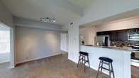 This single-floor space showcases an open-concept design that integrates the living area and kitchen into one cohesive layout. The living area is finished with wall-to-wall carpeting in a neutral tone and painted in a soft blue-gray hue, creating a calm and welcoming atmosphere. A modern track lighting fixture mounted on the ceiling provides focused illumination across the room. A hallway visible in the background leads to additional areas of the unit, adding a sense of depth to the floor plan.

The kitchen is separated from the living space by a partial wall that forms a breakfast bar, topped with a light granite-style countertop and accommodating two bar stools — an ideal setup for casual dining or entertaining. Dark espresso cabinetry contrasts cleanly against the lighter walls and countertops, while stainless steel appliances, including a refrigerator, contribute a contemporary finish. A window with horizontal blinds allows natural light to filter into the kitchen. A small floral arrangement on the counter adds a subtle decorative touch. Overall, the space presents a functional and modern interior with a consistent, neutral color palette throughout.