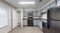 This single-floor residence offers a thoughtfully laid-out living space with a mix of practical finishes and comfortable design throughout. The kitchen serves as a functional centerpiece, featuring gray cabinetry, dark granite-style countertops, and a full suite of black appliances including a range with overhead microwave, dishwasher, and refrigerator. A dedicated walk-in pantry with open shelving provides generous storage, while light wood-look flooring ties the space together cleanly. The living area welcomes natural light through a large window dressed with blinds, and features the same durable hardwood-style flooring that extends through the main areas of the home. Bedrooms are carpeted for added comfort and each includes a ceiling fan with light fixture and a window that allows for natural light. The textured ceilings are consistent throughout the home, adding a classic residential feel. The bathroom is equipped with a full tub-shower combo featuring tile surround, a vanity with matching granite-style countertop, and hardwood-style flooring. The overall layout, as reflected in the dollhouse view, flows efficiently with well-defined zones for living, cooking, and sleeping, making this a practical and comfortable single-level residence.