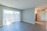 This single-floor residence offers a well-organized layout with distinct living, sleeping, kitchen, and bathroom areas. The living room serves as the central gathering space, finished with dark wood-style flooring and neutral taupe walls beneath a textured popcorn ceiling. A full-width sliding glass door dominates one wall, fitted with vertical blinds and opening to an outdoor patio area, allowing generous natural light to fill the room. The adjacent kitchen follows a galley configuration, featuring white upper cabinetry, grey lower cabinets, and crisp white countertops. Stainless steel appliances, including a refrigerator and an electric range, complement the functional design, while a window at the far end provides additional natural light and ventilation. The bedroom is finished with soft wall-to-wall carpeting, warm neutral walls, a textured ceiling, and a window with blinds that introduces natural light. A closet with a white paneled door provides practical storage. The bathroom is neatly appointed with a full soaking tub and shower combination, a curved chrome curtain rod, a vanity with white countertop, and light wood-style flooring, completing the unit's clean and cohesive aesthetic.