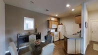 This single-story residence at Glenmary Village offers an open-concept layout that seamlessly connects the kitchen and dining areas, creating a cohesive and functional living space. The kitchen is equipped with white appliances, including a range, built-in microwave, and full-size refrigerator, all complemented by warm wood-toned cabinetry. A dark-finished kitchen island with a sink and contrasting countertop provides additional prep space and serves as a natural dividing point between the kitchen and dining areas. Rich dark hardwood-style flooring runs throughout, adding warmth and visual continuity to the space. The dining area accommodates a glass-top table with upholstered chairs, positioned alongside a decorative console and a framed wall mirror that enhances the sense of openness. Recessed lighting is installed throughout the ceiling, providing consistent, even illumination across the space. Neutral taupe wall tones contribute to a calm and inviting ambiance. A doorway visible from the kitchen suggests additional rooms beyond the main open area, rounding out this well-appointed single-floor home.