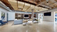 Located at 470 Market Ave SW in Grand Rapids, Michigan, this thoughtfully renovated single-floor space blends industrial heritage with modern design across a generously sized footprint. Exposed heavy timber beams, wood plank ceilings, and visible ductwork pay homage to the building's industrial roots, while polished concrete floors and crisp white walls create a clean, contemporary atmosphere throughout.

The main living and lounge area is an open, airy expanse anchored by a striking honeycomb-patterned wood feature wall with an integrated display screen. Large grid-pane windows flood the space with natural light, and a mix of seating arrangements—including plush yellow sofas, round tables with amber chairs, and bar-height seating—offer flexible social and work configurations. A black metal planter with live greenery adds a natural focal point to the open floor plan.

A dedicated collaborative zone features carpet-tiled flooring, bold geometric wallcovering, pendant lighting, and a glass-enclosed phone booth for private calls. A beverage nook with tile backsplash and cabinetry provides a convenient refreshment station nearby.

The well-appointed exercise room is a standout amenity, offering rubber flooring, full-wall mirrors, a comprehensive selection of cardio machines and strength equipment, and abundant natural light from oversized industrial windows. The hallway and supporting rooms connect the areas seamlessly, completing this versatile and dynamic space.