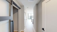 This contemporary single-floor residence showcases a clean, modern design with light hardwood flooring and a neutral gray-and-white palette throughout. A welcoming hallway with a sliding barn-style door provides a stylish entry, leading past storage and closet space into the main living area. The kitchen is a standout feature, outfitted with textured wood-grain cabinetry, matte black hardware, a white subway tile backsplash with under-cabinet lighting, light quartz countertops, and a full suite of stainless steel appliances including a refrigerator, range, microwave, and dishwasher. A freestanding kitchen island offers additional workspace and casual seating. The open layout connects seamlessly to the living area, which is bathed in natural light from expansive floor-to-ceiling windows framing panoramic city views. The bathroom is well-appointed with large-format tile flooring, a soaking tub and shower combo with full tile surround, a wood-finish vanity with a light countertop, and direct access to a walk-in closet with wire shelving. The space combines functional design with upscale finishes, creating a bright and cohesive modern living environment.