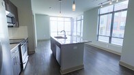 This single-floor space showcases a clean, contemporary design centered around an open-concept layout that seamlessly connects the kitchen and living areas. The kitchen island serves as the focal point, featuring a sleek stone countertop with an integrated sink and modern faucet, complemented by two stylish pendant lights suspended overhead. The island's neutral tones coordinate with the soft grey-green walls throughout the space.

Large floor-to-ceiling windows line multiple walls, fitted with horizontal blinds that allow for adjustable natural light. This abundance of glazing creates a bright, airy atmosphere and offers views of the surrounding exterior. Dark hardwood flooring runs throughout, providing a rich contrast to the light-toned walls and cabinetry, adding warmth and visual depth to the overall aesthetic.

The open floor plan allows for a versatile arrangement of the living and dining areas, which flow naturally from the kitchen. Track lighting along the ceiling supplements the natural light. The minimalist design approach, clean lines, and neutral color palette give the space a modern, functional character suited to contemporary living.