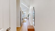 This efficiently designed one-bedroom, one-bathroom unit in Russellville, Arkansas makes smart use of its compact footprint through thoughtful layout and striking architectural details. The most notable feature is the dramatically vaulted ceiling that rises over the main living and kitchen corridor, lending an airy, open quality far beyond what the floor area might suggest. Sleek linear pendant lighting runs along this vaulted ceiling, further emphasizing the vertical space.

The galley-style kitchen lines one wall with light wood cabinetry, white countertops, stainless steel appliances including a refrigerator, microwave, and range, and open upper shelving for additional storage. Light wood-tone flooring runs continuously throughout the unit, unifying each space cohesively.

The living area benefits from a large picture window that frames tree-lined outdoor views and floods the room with natural light. A wall-mounted television and compact dining table make this space both functional and comfortable.

The bedroom, though cozy in scale, is well-proportioned and features the same warm flooring and bright white walls. The bathroom includes a shower with curtain, open shelving, and a stacked washer and dryer unit, efficiently combining laundry and bath functions within a single, well-organized room.