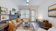 This single-floor residence showcases a thoughtfully designed open-concept layout with contemporary finishes throughout. The living area features light wood-look flooring, a distinctive dark shiplap accent wall with a display ledge, and direct access to an exterior door alongside generous windows that fill the space with natural light. The kitchen is a standout, equipped with flat-front light wood cabinetry extending to the ceiling, granite-style countertops, a subway tile backsplash with under-cabinet lighting, stainless steel appliances, and a central island with a undermount sink and seating — ideal for both cooking and casual dining. Pendant lighting adds a modern finishing touch overhead. The bedroom offers a calm, neutral palette with sage-toned walls, large windows with outdoor views, and light hardwood flooring. The bathroom is spacious and well-appointed, featuring a walk-in glass shower, an extended vanity with granite countertops, light wood cabinetry, open wooden shelving, and coordinating black hardware throughout. A walk-in closet with wire shelving provides practical storage. Together, these spaces create a cohesive, modern aesthetic that balances functionality with refined design.