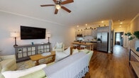 This single-floor home offers a thoughtfully designed open-concept layout that seamlessly connects the living, dining, and kitchen areas. Rich hardwood flooring flows throughout, anchoring the warm and inviting atmosphere.

The living area is bright and welcoming, featuring crown molding, a ceiling fan, and large windows dressed with floral-patterned curtains that invite generous natural light. Comfortable seating, a patterned area rug, and a wall-mounted flat-screen TV complete the relaxed yet polished look.

The kitchen is both functional and stylish, showcasing gray shaker-style cabinetry extending to the ceiling, a herringbone tile backsplash, stainless steel appliances including a side-by-side refrigerator with ice maker, a gas range, and a built-in microwave. A butcher block dining table with bar-style seating serves as the central gathering point between the kitchen and living space.

The bedroom is a tranquil retreat with soft gray walls, crown molding, a ceiling fan, and windows framed by elegant sage green drapes. A powder blue upholstered headboard and coordinating bedding add a coastal charm.

The bathroom features light hardwood flooring, a gray vanity, a tall linen cabinet, and a walk-in shower with dark subway tile, creating a clean and modern aesthetic.