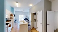 This single-floor residence offers a thoughtfully designed open-concept layout that seamlessly connects the kitchen, dining, and living areas. The kitchen is anchored by a white island with bar-style seating, providing both a functional prep space and a casual dining spot. A stainless steel sink with a modern gooseneck faucet adds a contemporary touch, while the refrigerator is conveniently positioned nearby. Light wood-tone flooring flows throughout the space, enhancing the bright and airy atmosphere created by the abundant natural light. A large sliding glass door or window in the living area frames an outdoor view and fills the room with daylight. The living area is generously proportioned, with enough space to accommodate a full sofa arrangement and a ceiling fan overhead for added comfort. Just off the main living space, a dedicated in-unit laundry closet houses a full stacked washer and dryer unit, offering exceptional convenience. The clean white walls and modern flush-mount lighting throughout contribute to a cohesive, neutral aesthetic. This well-appointed space is designed for comfortable, efficient everyday living with a modern sensibility.