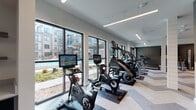 This contemporary fitness center is thoughtfully designed to support a full range of workout routines in a bright, stylish environment. A defining feature of the space is the floor-to-ceiling windows lining one wall, which flood the interior with natural light and offer direct views of an outdoor pool courtyard, creating an energizing atmosphere for exercise.

The cardio zone is lined with premium equipment, including interactive cycling bikes with integrated display screens, StairMaster steppers, elliptical machines, and multiple treadmills — all positioned to take advantage of the outdoor views. The flooring throughout features a striking chevron-patterned tile that adds visual interest and defines the open layout.

The strength training area is equally well-appointed, offering a full dumbbell rack, weight benches, cable and functional training machines, and stability balls. Full-length mirrors in the strength zone enhance the sense of space and allow for proper form monitoring. A bold geometric accent wall in teal and charcoal tones adds a dynamic, modern aesthetic to this section of the gym.

Overhead, linear LED lighting runs the length of the ceiling, providing consistent, even illumination throughout. Structural columns with white shiplap detailing contribute architectural character while subtly dividing the cardio and strength zones. The overall design is clean, modern, and purpose-built for an active lifestyle.