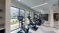 This contemporary fitness center is thoughtfully designed to support a full range of workout routines in a bright, stylish environment. A defining feature of the space is the floor-to-ceiling windows lining one wall, which flood the interior with natural light and offer direct views of an outdoor pool courtyard, creating an energizing atmosphere for exercise.

The cardio zone is lined with premium equipment, including interactive cycling bikes with integrated display screens, StairMaster steppers, elliptical machines, and multiple treadmills — all positioned to take advantage of the outdoor views. The flooring throughout features a striking chevron-patterned tile that adds visual interest and defines the open layout.

The strength training area is equally well-appointed, offering a full dumbbell rack, weight benches, cable and functional training machines, and stability balls. Full-length mirrors in the strength zone enhance the sense of space and allow for proper form monitoring. A bold geometric accent wall in teal and charcoal tones adds a dynamic, modern aesthetic to this section of the gym.

Overhead, linear LED lighting runs the length of the ceiling, providing consistent, even illumination throughout. Structural columns with white shiplap detailing contribute architectural character while subtly dividing the cardio and strength zones. The overall design is clean, modern, and purpose-built for an active lifestyle.