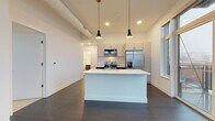 Located at 230 W Cherry St in Milwaukee's vibrant 53212 zip code, this contemporary single-floor unit showcases a thoughtfully designed open-concept layout. The living and kitchen area is anchored by a large white center island with a built-in sink, complemented by pendant lighting and exposed ductwork that adds an industrial-modern character. Light gray cabinetry lines the kitchen walls, paired with a full suite of stainless steel appliances including a French door refrigerator, electric range, and over-the-range microwave. Dark hardwood-style flooring extends throughout the main living area, grounding the bright, white interior. Floor-to-ceiling sliding glass doors open to a private balcony, flooding the space with natural light and offering open city views. The bedroom features plush carpeting and tall, narrow windows that frame additional urban views of the Milwaukee streetscape. A walk-in closet with wire shelving systems provides generous storage. The bathrooms are finished cleanly in white, one featuring a full soaking tub and shower combination, the other a walk-in shower with a floating dark-cabineted vanity and large mirror. Light tile flooring completes the bathrooms' crisp, modern aesthetic throughout.