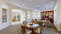 This thoughtfully designed single-floor space offers a warm and inviting atmosphere ideal for gathering and relaxation. The main common area serves as the heart of the space, featuring a round wooden dining table surrounded by cushioned chairs, and a plush seating arrangement with a sofa and accent chairs. Arched doorways lend an architectural elegance that connects the various areas seamlessly, while recessed lighting and large windows fitted with wooden blinds flood the interior with generous natural light and views of lush greenery outside.

A standout feature of the living area is the rich, dark wood built-in entertainment center, complete with open display shelves showcasing decorative accents, ceramic vases, and curated collectibles, alongside a centrally mounted television and louvered cabinet doors below. A patterned accent chair and a glass-top wrought iron coffee table complete this cozy corner.

The kitchenette is efficiently laid out with dark granite countertops, a peninsula with a sink, and warm cherry wood cabinetry. Light tile flooring grounds the space, contrasting with the neutral carpeting found throughout the main areas. Together, these spaces create a cohesive, comfortable environment with a classic and refined aesthetic suitable for community or shared use.