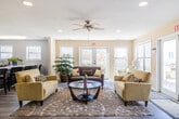 This single-floor community clubhouse offers a thoughtfully designed layout that seamlessly connects multiple functional areas, creating an inviting and practical environment for residents. The main lounge serves as the heart of the space, featuring light hardwood-style flooring, a textured ceiling with a ceiling fan, and an abundance of natural light streaming through multiple large windows and French doors that open to an outdoor area. Comfortable seating arrangements — including warm yellow sofas and a dark upholstered couch centered around a glass-top coffee table — give the lounge a welcoming, social atmosphere. Adjacent to the lounge, French-paned glass doors lead into a secondary sitting area that flows into the leasing office. The office is well-appointed with a rich wood reception desk, accent seating in chartreuse upholstery, and direct access to an exterior entrance. The kitchen is a standout feature, equipped with dark espresso cabinetry, stainless steel appliances, light countertops, and a large island with bar seating — ideal for communal gatherings. A dining area and an art-lined corridor connect the spaces, with framed artwork adding character throughout. The overall ambiance is bright, clean, and welcoming.