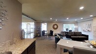 This single-floor space is thoughtfully arranged to accommodate a variety of activities within one open, flowing layout. At the forefront, a dark wood counter with a granite surface serves as a welcoming reception or service area. A glass partition wall on the left side adds a modern architectural element, visually separating an adjacent space while maintaining an open, connected feel throughout.

The main area is divided into several functional zones. A glass-top dining or meeting table surrounded by dark chairs provides a spot for collaborative gatherings, while a dedicated workstation area near the windows is equipped with computers, catering to productivity needs. A comfortable seating lounge with tufted black chairs and a glass coffee table offers a relaxed environment for informal conversations or leisure.

Recessed ceiling lighting illuminates the neutral-toned walls and carpeted flooring, creating a warm and professional ambiance. Natural light filters in through multiple windows, enhancing the brightness of the space. Decorative touches, including a round sunburst mirror and a bookshelf, add character and a sense of comfort to this well-appointed, multi-use space.