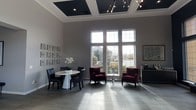 This refined single-floor space makes a striking impression through its thoughtful architectural design and carefully curated interior. The room commands attention with its dramatically high ceilings, accented by bold dark coffered panels that contrast elegantly against the bright white crown molding. A sleek, multi-arm chandelier with exposed bulbs adds a contemporary edge overhead, complementing the space's modern-classic aesthetic.

Tall, multi-pane windows dominate one wall, flooding the interior with natural light and offering open views of the surrounding landscaped exterior. The soft gray tile flooring runs throughout, providing a clean, neutral foundation that ties the design elements together cohesively.

The space is furnished with a mix of seating arrangements that balance functionality and style. A round white pedestal table paired with dark leather chairs anchors one area, while a pair of plush deep-burgundy armchairs and a delicate glass side table create an inviting conversational grouping nearby. Decorative wall art in a grid arrangement adds visual interest to the neutral gray walls, alongside framed artwork that reinforces the polished ambiance.

Overall, this space exudes a professional yet welcoming atmosphere, ideal for reception, meeting, or lounge purposes, with its blend of elevated design details and comfortable furnishings.