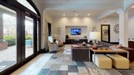 Located at 300 Blue Moon Crossing in Pooler, Georgia, this single-floor community space offers an open, airy layout designed for both comfort and functionality. The interior is defined by soaring ceilings, recessed lighting, and a sophisticated palette of warm neutrals accented by rich, dark wood finishes.

The expansive lounge area features multicolored natural stone tile flooring that adds texture and visual interest throughout. Generous seating arrangements, including plush sofas and accent chairs, are organized around a wall-mounted television set within a built-in media niche. Arched architectural details and large floor-to-ceiling glass doors flood the space with natural light and provide views of the outdoor patio area.

The gourmet kitchen is a standout feature, showcasing dark espresso cabinetry, granite countertops, and a large central island topped with matching stone surfaces. A bold stainless steel column range hood rises dramatically above the island's built-in cooktop, serving as an architectural focal point. Stainless steel appliances, including a double wall oven and French door refrigerator, complete the professional-grade setup. A granite-topped breakfast bar with stylish barstool seating offers an additional gathering spot.

A dedicated dining and work area rounds out the space, providing versatile seating for meetings or casual dining. The overall ambiance is modern, welcoming, and well-appointed.