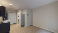 This single-floor studio unit offers a clean, modern layout designed for efficient use of space. The main living area features light wood-tone laminate flooring and neutral warm-gray walls, creating a bright and welcoming atmosphere. A wall-mounted thermostat and control panel are conveniently positioned within the living space. Large windows along one wall invite generous natural light and provide views of the surrounding building exterior, enhancing the sense of openness within the unit.

The kitchenette is outfitted with dark espresso-toned cabinetry, both upper and lower, paired with a light countertop and a stainless steel sink. Black appliances, including a microwave, are integrated seamlessly into the cabinetry, offering a sleek and functional cooking area.

The bathroom is accessible directly from the main living space and includes a full shower and tub combination with chrome fixtures, a toilet, and a towel bar. The bathroom features light-toned flooring and neutral white walls, maintaining the unit's clean and uniform aesthetic.

The entry area includes a solid white paneled front door alongside an interior door providing access to the bathroom, establishing a clear and practical flow throughout the unit. Overall, the space presents a well-organized, move-in-ready layout with consistent modern finishes throughout.