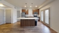 This contemporary single-floor space is thoughtfully designed with a clean, modern aesthetic throughout. The heart of the unit is an open-concept kitchen and living area, where rich walnut-toned cabinetry pairs elegantly with bright white quartz countertops. A central kitchen island with pendant lighting above serves as both a functional workspace and a natural gathering point. The kitchen is fully equipped with stainless steel appliances, including a refrigerator, range, microwave, and dishwasher, all set against a classic white subway tile backsplash. A deep single-basin sink with a flexible faucet adds practical functionality. Durable wood-look flooring flows throughout the main living and kitchen areas, lending warmth and continuity to the space. Natural light enters through glass patio doors, brightening the open layout. The bathroom continues the design language of the kitchen, featuring matching walnut cabinetry, white quartz countertops, a large vanity mirror, and chrome fixtures. The bedroom offers a quieter retreat with plush carpeting underfoot and a window that brings in natural light. Overall, the space presents a cohesive, contemporary feel with consistent finishes and a practical, well-organized layout throughout.