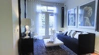 This single-floor living space showcases a carefully curated interior defined by a striking navy and white color palette. The living area serves as the centerpiece, anchored by a deep navy sofa adorned with patterned and textured throw pillows that add visual depth. A sleek white coffee table with a chrome frame sits atop a dark, speckled area rug, creating an elegant contrast against the lighter surroundings.

Natural light fills the room through large French doors dressed in sheer white curtains, which open to an outdoor space framed by lush greenery. The doors feature transom windows above, further enhancing the sense of openness and brightness. A dramatic pendant light fixture in black adds a vertical design element, complementing the room's bold aesthetic.

Decorative accents include classic white candlestick holders of varying heights, a sculptural white table lamp with a dark base, and a framed monochromatic artwork on the wall. A dark-stained console table near the entrance adds a refined, formal touch. Together, these elements create a sophisticated, design-forward space that balances contrast and elegance throughout.