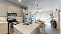 This single-floor residence showcases a thoughtfully designed open-concept layout that seamlessly connects the kitchen, dining, and living areas. The kitchen is a standout feature, equipped with stainless steel appliances, light wood cabinetry extending to the ceiling, and a generously sized island topped with quartz countertops. The island doubles as a breakfast bar with seating, while a pendant light fixture adds a decorative focal point. Recessed lighting and wood-look flooring enhance the space throughout.

The living area benefits from large floor-to-ceiling windows fitted with roller shades, allowing abundant natural light to filter in. A ceiling fan adds functional comfort to the space.

The primary bedroom offers plush carpeting, a ceiling fan, and large windows with roller shades and curtain panels, creating a calm, restful retreat. A secondary bedroom features a similar neutral palette with ample natural light.

The primary bathroom includes a glass-enclosed shower, a floating vanity with quartz countertops, a large mirror, and direct access to a walk-in closet with built-in storage. A powder room features a bold patterned accent wall, light wood vanity, and quartz countertop, adding a stylish contrast to the otherwise neutral palette throughout the residence.