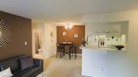 This single-floor space is designed around an open-concept layout that integrates the living, dining, and kitchen areas into one cohesive and functional environment. The living area features plush wall-to-wall carpeting and accommodates comfortable seating, creating an inviting space for relaxation. A half-wall peninsula separates the kitchen from the rest of the space, providing both a visual boundary and a practical surface for casual dining or food preparation.

The dining area is defined by a rich, warm brown accent wall that serves as a focal point, adding depth and character to the interior. A ceiling fan with a light fixture overhead provides both ventilation and ambient lighting to this zone. A compact bistro-style dining set with bar-height chairs fits neatly within the space.

The kitchen is positioned adjacent to the dining area, with the peninsula offering additional counter space. A bathroom is visible through a doorway at the rear of the space, featuring a shower-tub combination and standard fixtures. The overall color palette consists of neutral tones with warm brown accents, creating a cozy and welcoming atmosphere throughout.