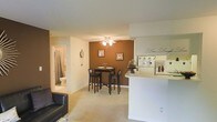 This single-floor space is designed around an open-concept layout that integrates the living, dining, and kitchen areas into one cohesive and functional environment. The living area features plush wall-to-wall carpeting and accommodates comfortable seating, creating an inviting space for relaxation. A half-wall peninsula separates the kitchen from the rest of the space, providing both a visual boundary and a practical surface for casual dining or food preparation.

The dining area is defined by a rich, warm brown accent wall that serves as a focal point, adding depth and character to the interior. A ceiling fan with a light fixture overhead provides both ventilation and ambient lighting to this zone. A compact bistro-style dining set with bar-height chairs fits neatly within the space.

The kitchen is positioned adjacent to the dining area, with the peninsula offering additional counter space. A bathroom is visible through a doorway at the rear of the space, featuring a shower-tub combination and standard fixtures. The overall color palette consists of neutral tones with warm brown accents, creating a cozy and welcoming atmosphere throughout.